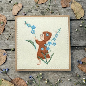 May include: A square greeting card featuring an embroidered design. The design depicts a child in a brown hooded outfit reaching for blue forget-me-not flowers. The card has a cream-colored felt background and a brown border.