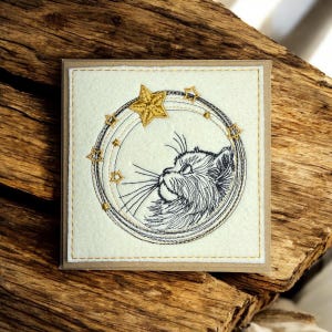 Op de afbeelding: Vierkante wenskaart met een crème vilt voorkant. Het heeft een geborduurde kattenkop in een cirkelvormig frame, met gouden sterren en een grote ster. De kaart heeft een bruine rand en gouden stiksels.