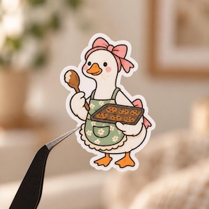 May include: A die-cut sticker featuring a white goose wearing a pink bow and a sage green apron with a white daisy pattern. The goose holds a wooden spoon in one wing and a dark grey baking sheet with six brown chocolate chip cookies in the other.
