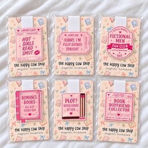 Pode incluir: Seis marcadores de página magnéticos em embalagens plásticas transparentes, cada um em um cartão floral com a etiqueta the happy cow shop. Os designs incluem cartões de biblioteca rosa, um bilhete, um distintivo, um livro e uma caixa de fósforos.