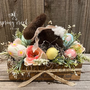 May include: An Easter display in a wooden crate, featuring a brown bunny, pastel eggs, and artificial flowers. The crate is tied with a ribbon and has text that reads "CHEESE" and "CREAMERY CO.". The arrangement is set against a rustic wooden background.