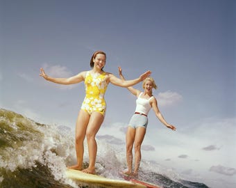 May include: Two women in swimsuits are riding a surfboard on a wave. The woman at the front is wearing a yellow and white floral swimsuit and the woman at the back is wearing a white tank top and blue shorts.