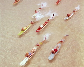 May include: A group of people are surfing on colourful surfboards in a shallow body of water. The water is clear and the sand is visible at the bottom. The people are wearing swimsuits and are smiling.