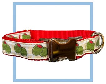 Puede incluir: Collar de perro rojo con una cinta estampada en blanco y verde con aceitunas. El collar tiene una hebilla plateada y un anillo en D plateado.