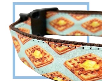 Puede incluir: Un collar de perro marrón con fondo azul claro y un patrón de galletas amarillas y naranjas. El collar tiene una hebilla de plástico negra.