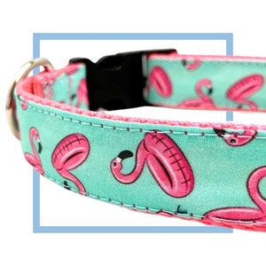 May include: A turquoise blue dog collar with pink flamingo print. The collar has a black plastic buckle and a silver D-ring.