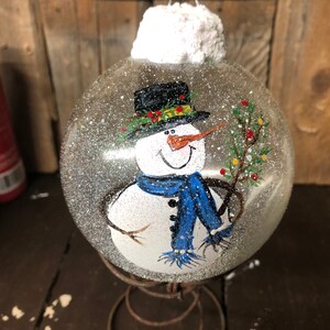 May include: A clear glass Christmas ornament with a hand-painted snowman wearing a black hat, blue scarf, and holding a green sprig. The ornament is covered in white glitter and has a white fluffy top.