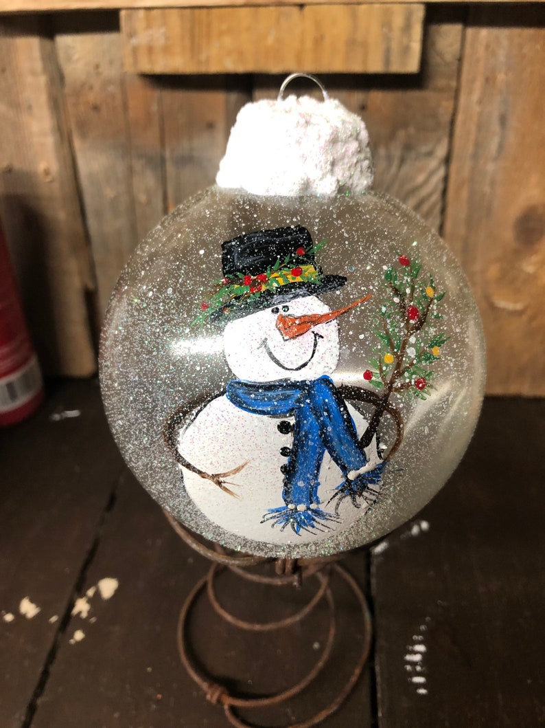 May include: A clear glass Christmas ornament with a hand-painted snowman wearing a black hat, blue scarf, and holding a green sprig. The ornament is covered in white glitter and has a white fluffy top.