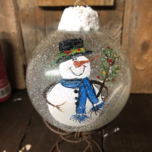 May include: A clear glass Christmas ornament with a hand-painted snowman holding a small green tree. The snowman is wearing a black top hat with a green and yellow trim, a blue scarf, and a white hat. The ornament is covered in white glitter.