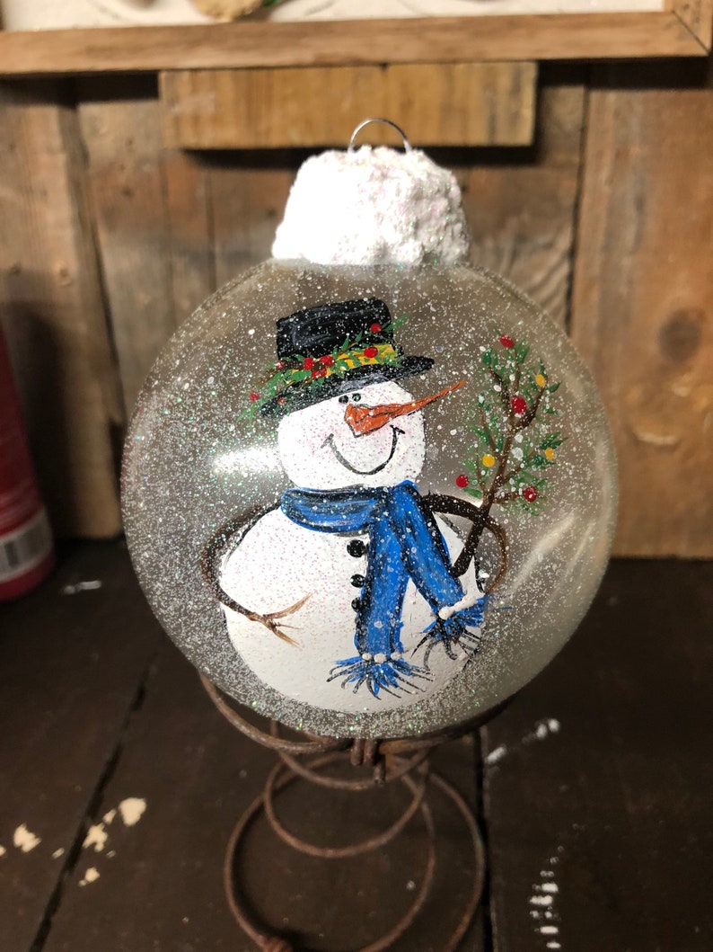 May include: A clear glass Christmas ornament with a hand-painted snowman holding a small green tree. The snowman is wearing a black top hat with a green and yellow trim, a blue scarf, and a white hat. The ornament is covered in white glitter.