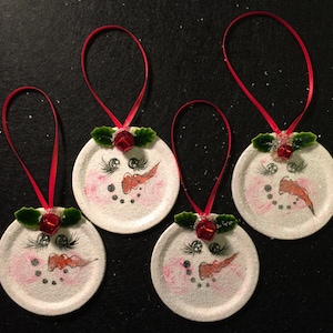 May include: Four white round ornaments with painted snowman faces, red bows, and green holly leaves. Each ornament has a red ribbon for hanging.
