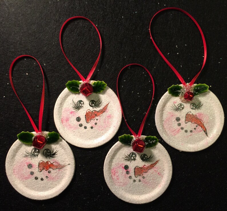 May include: Four white round ornaments with painted snowman faces, red bows, and green holly leaves. Each ornament has a red ribbon for hanging.