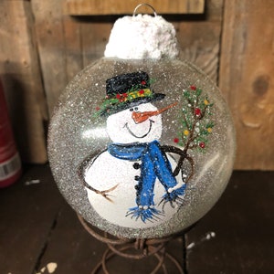 May include: A clear glass Christmas ornament with a hand-painted snowman wearing a black hat, blue scarf, and holding a twig with red berries. The ornament is covered in white glitter to resemble snow.
