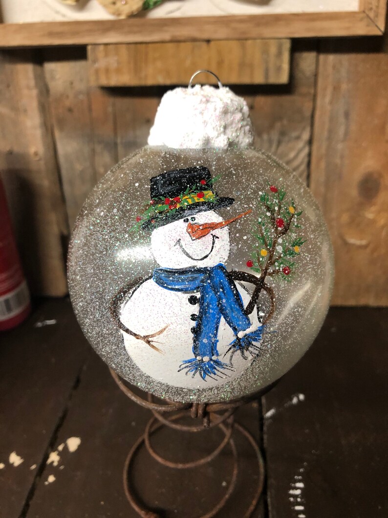 May include: A clear glass Christmas ornament with a hand-painted snowman wearing a black hat, blue scarf, and holding a twig with red berries. The ornament is covered in white glitter to resemble snow.