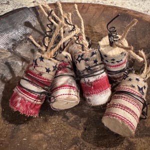 May include: A collection of six patriotic-themed ornaments. Each ornament is cylindrical, wrapped in fabric with red and white stripes and a blue star pattern. They are tied with wire and twine, resembling miniature firecrackers.