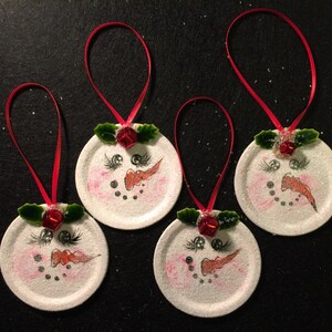 May include: Four white snowman ornaments with red ribbon, red noses, black eyes, and green holly leaves. The ornaments are made from repurposed jar lids.