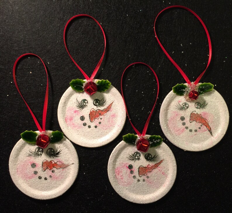 May include: Four white snowman ornaments with red ribbon, red noses, black eyes, and green holly leaves. The ornaments are made from repurposed jar lids.