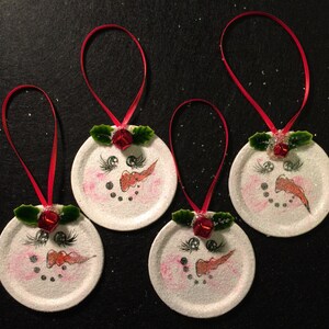 May include: Four white snowman ornaments with red ribbon hangers. Each ornament has a painted snowman face with black eyes, a red nose, and a black mouth. The snowmen have green holly leaves and a red bell on top.