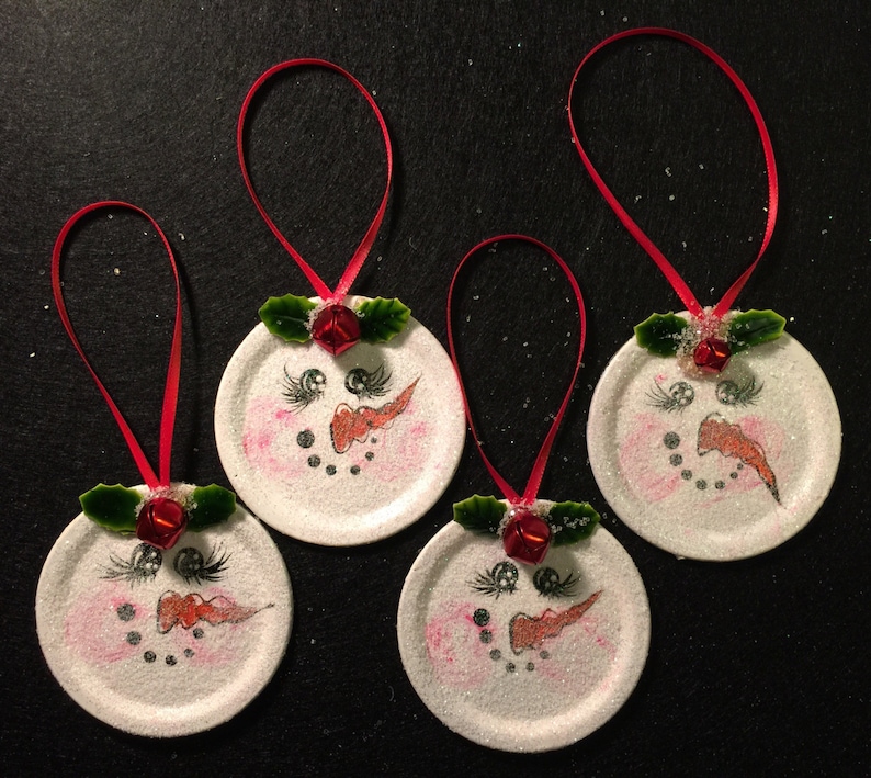 May include: Four white snowman ornaments with red ribbon hangers. Each ornament has a painted snowman face with black eyes, a red nose, and a black mouth. The snowmen have green holly leaves and a red bell on top.