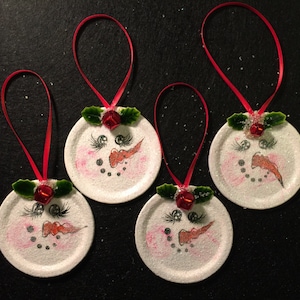 May include: Four white snowman ornaments with red ribbon hangers. Each ornament has a painted snowman face with black eyes, a red nose, and a black mouth. Each ornament has a green holly sprig with a red bell.