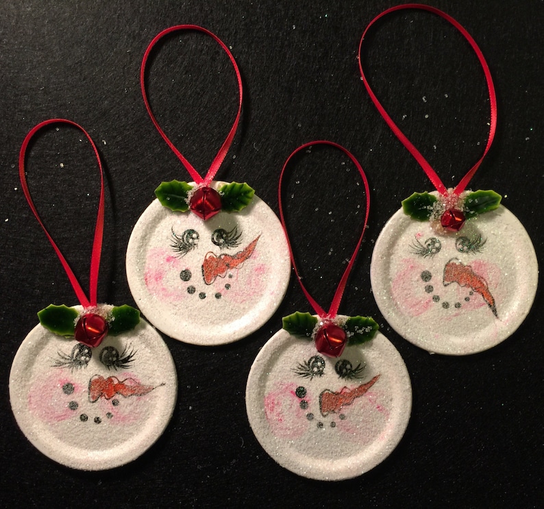 May include: Four white snowman ornaments with red ribbon hangers. Each ornament has a painted snowman face with black eyes, a red nose, and a black mouth. Each ornament has a green holly sprig with a red bell.