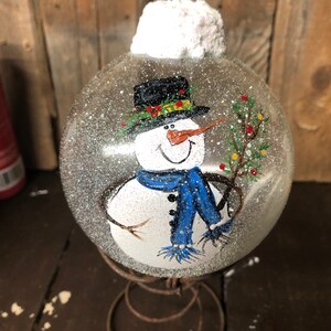 May include: A hand-painted glass ornament featuring a snowman with a black top hat, blue scarf, and a twig in its hand. The ornament is covered in glitter and has a white fluffy top.