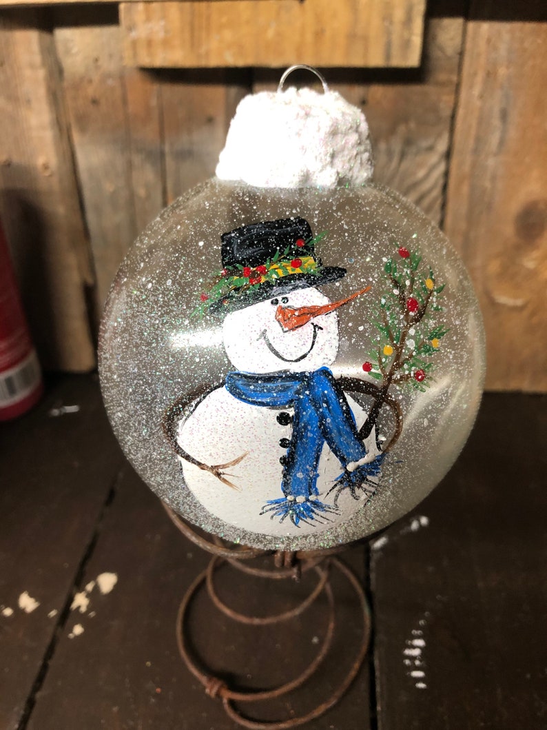 May include: A hand-painted glass ornament featuring a snowman with a black top hat, blue scarf, and a twig in its hand. The ornament is covered in glitter and has a white fluffy top.