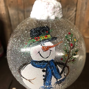 May include: A clear glass Christmas ornament with a hand-painted snowman design. The snowman is wearing a black top hat with a green and red trim, a blue scarf, and holding a small green branch with red berries. The ornament is covered in white glitter to resemble snow.