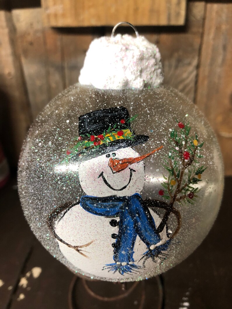 May include: A clear glass Christmas ornament with a hand-painted snowman design. The snowman is wearing a black top hat with a green and red trim, a blue scarf, and holding a small green branch with red berries. The ornament is covered in white glitter to resemble snow.