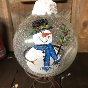 May include: A hand-painted glass ornament featuring a snowman with a black top hat, blue scarf, and a twig in its hand. The ornament is covered in white glitter and has a white felt top.