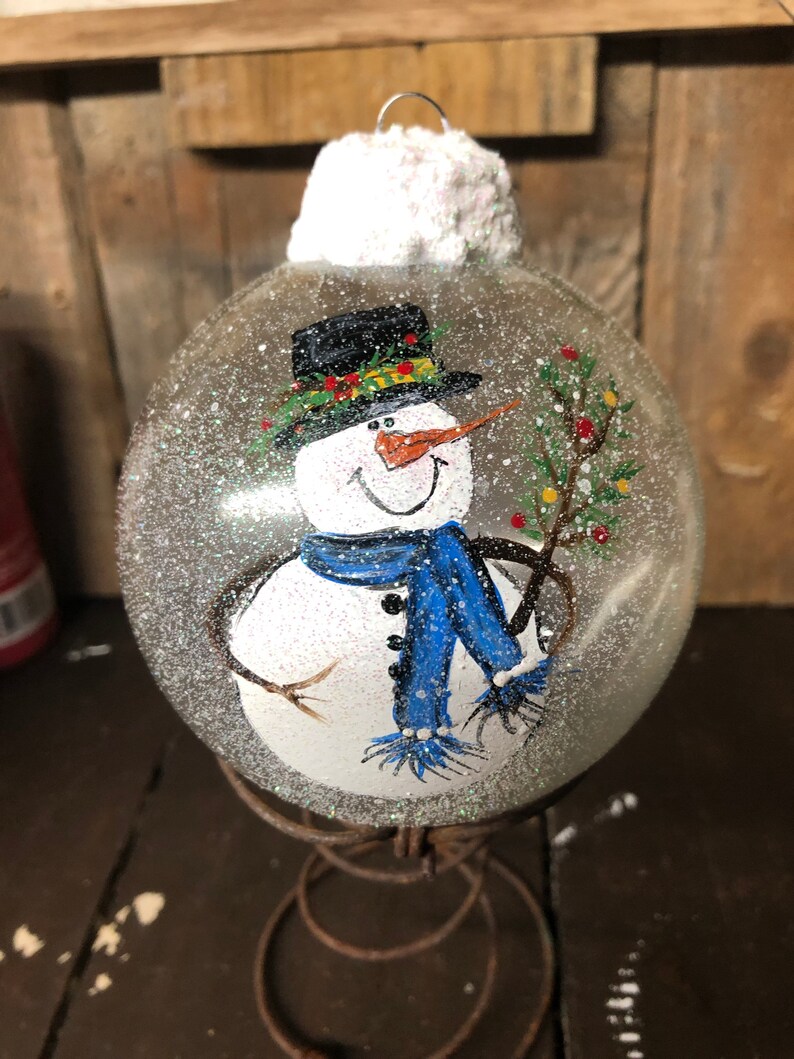 May include: A hand-painted glass ornament featuring a snowman with a black top hat, blue scarf, and a twig in its hand. The ornament is covered in white glitter and has a white felt top.