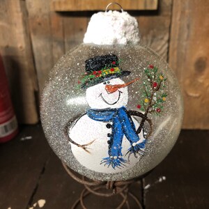 May include: A clear glass Christmas ornament with a hand-painted snowman wearing a black top hat, blue scarf, and holding a twig with green leaves. The ornament is covered in white glitter and has a white fluffy top. The ornament is sitting on a rusty metal stand.