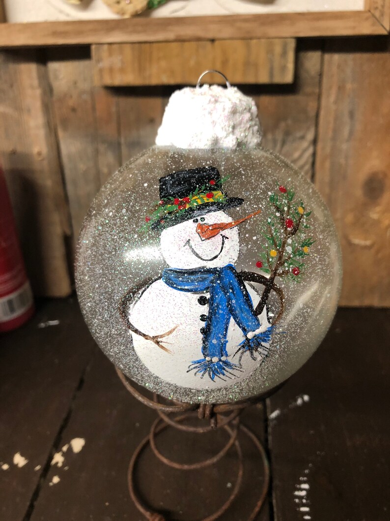 May include: A clear glass Christmas ornament with a hand-painted snowman wearing a black top hat, blue scarf, and holding a twig with green leaves. The ornament is covered in white glitter and has a white fluffy top. The ornament is sitting on a rusty metal stand.