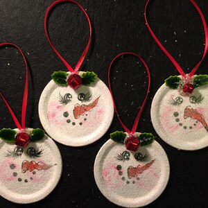 May include: Four white painted jar lid ornaments with red ribbon, green holly leaves, and red bells. Each ornament has a snowman face painted on it with black eyes, a red nose, and a black mouth.
