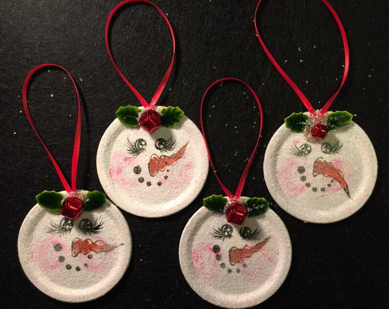 May include: Four white painted jar lid ornaments with red ribbon, green holly leaves, and red bells. Each ornament has a snowman face painted on it with black eyes, a red nose, and a black mouth.