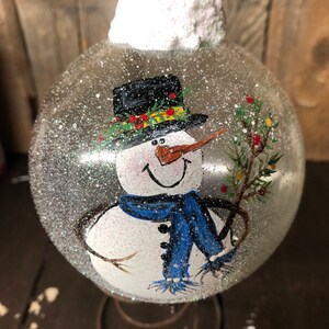 May include: A clear glass Christmas ornament with a painted snowman design. The snowman is wearing a black top hat with a green and yellow trim, a blue scarf, and a carrot nose. The ornament is covered in white glitter to resemble snow.