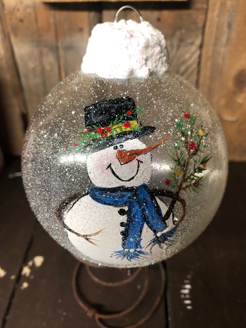 May include: A clear glass Christmas ornament with a painted snowman design. The snowman is wearing a black top hat with a green and yellow trim, a blue scarf, and a carrot nose. The ornament is covered in white glitter to resemble snow.