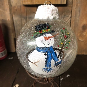 May include: A clear glass Christmas ornament with a painted snowman holding a small green tree. The snowman is wearing a black top hat, a green scarf, a blue scarf, and white mittens. The ornament has a white, fluffy top and is sitting on a rusty metal stand.