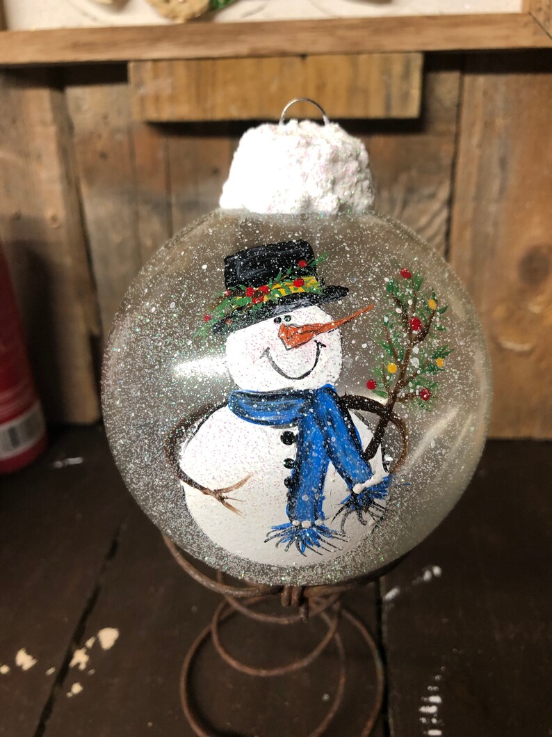May include: A clear glass Christmas ornament with a painted snowman holding a small green tree. The snowman is wearing a black top hat, a green scarf, a blue scarf, and white mittens. The ornament has a white, fluffy top and is sitting on a rusty metal stand.
