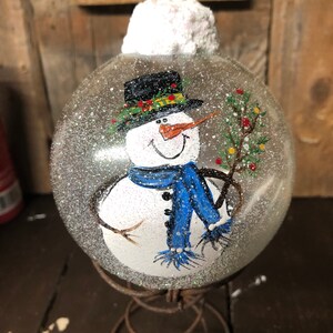 May include: A clear glass Christmas ornament with a hand-painted snowman wearing a black hat, blue scarf, and holding a branch with red berries. The ornament is covered in glitter and has a white fluffy top.