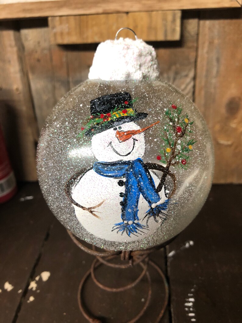 May include: A clear glass Christmas ornament with a hand-painted snowman wearing a black hat, blue scarf, and holding a branch with red berries. The ornament is covered in glitter and has a white fluffy top.