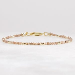 May include: Delicate beaded bracelet featuring small, faceted, light brown beads interspersed with gold-colored metal beads. The bracelet has a gold-colored clasp and is displayed on a white surface. A simple and elegant piece of jewelry.