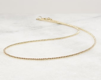 May include: A delicate gold chain necklace composed of small, uniform, round metallic beads. The necklace features a polished, reflective finish and includes a small circular clasp mechanism visible at the end of the chain, creating a continuous, refined look.