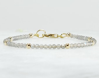 May include: A delicate bracelet featuring small, faceted grey labradorite beads interspersed with smooth, polished gold-tone metal spheres. The piece is secured with a gold-tone metal lobster claw clasp and a circular jump ring connector.