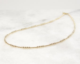 May include: A gold-toned beaded necklace featuring a continuous strand of small, polished, spherical beads of varying sizes arranged in a repeating pattern, secured with a small metal clasp at the back of the circular chain design.