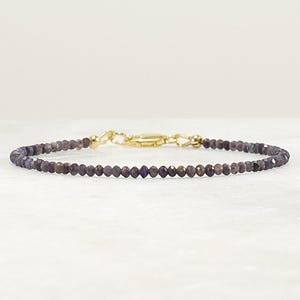 May include: A delicate bracelet composed of small, faceted dark purple beads with a reflective finish. The piece features a gold-toned metal lobster claw clasp and connecting hardware, creating a simple and refined circular design for the wrist.