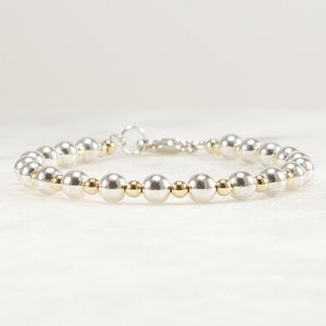May include: A beaded bracelet featuring alternating large silver-toned spheres and smaller gold-toned spheres. The bracelet is finished with a silver-toned lobster claw clasp and a circular jump ring, creating a continuous circular design.