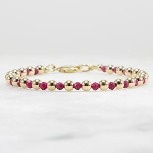 May include: Gold beaded bracelet with alternating ruby and gold beads. The bracelet features a gold clasp and is displayed on a white surface. The gold beads are round and polished, while the ruby beads are faceted.