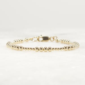 May include: A gold-toned beaded bracelet featuring a series of polished, spherical beads of varying sizes arranged in a repeating pattern, secured with a metallic lobster claw clasp mechanism at the center of the circular band design.