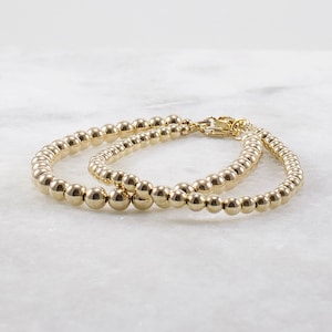 May include: Two gold beaded bracelets with a clasp closure. The bracelets are made of small, round beads.